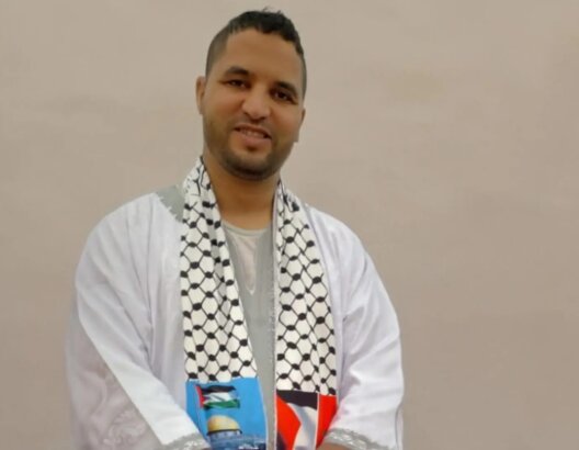 شخص يرتدي ثوباً أبيض ووشاحاً فلسطينياً يحمل كوباً أزرق في خلفية بسيطة