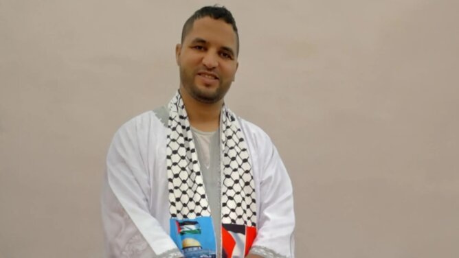 شخص يرتدي ثوباً أبيض ووشاحاً فلسطينياً يحمل كوباً أزرق في خلفية بسيطة