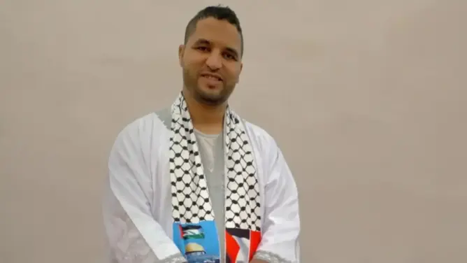 شخص يرتدي ثوباً أبيض ووشاحاً فلسطينياً يحمل كوباً أزرق في خلفية بسيطة