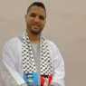 شخص يرتدي ثوباً أبيض ووشاحاً فلسطينياً يحمل كوباً أزرق في خلفية بسيطة
