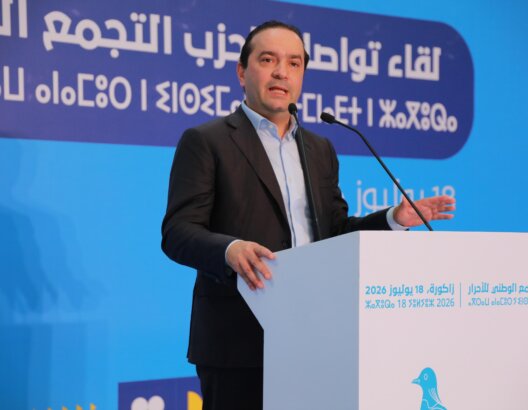 محمد شوكي رئيس حزب التجمع الوطني للأحرار يتحدث في اجتماع عام خلف منبر أبيض وشعار حزب التجمع الوطني للأحرار