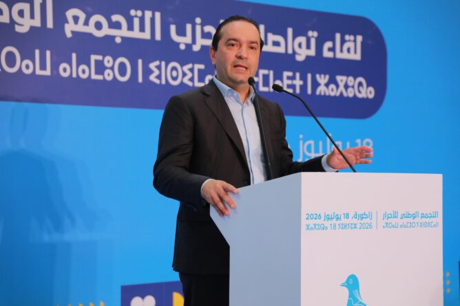 محمد شوكي رئيس حزب التجمع الوطني للأحرار يتحدث في اجتماع عام خلف منبر أبيض وشعار حزب التجمع الوطني للأحرار