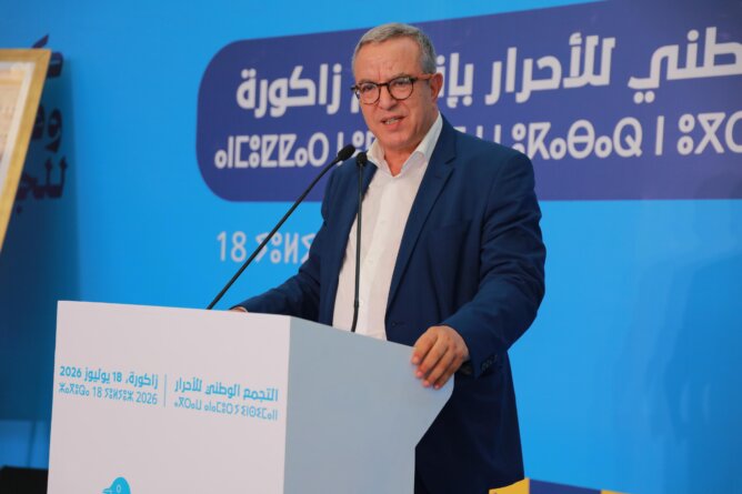 محمد أوجار خلف منبر أبيض في لقاء حزبي بخلفية زرقاء ونصوص عربية