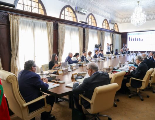 اجتماع رسمي في غرفة مزخرفة لمناقشة تقارير مالية مع علم المغرب وشاشة عرض