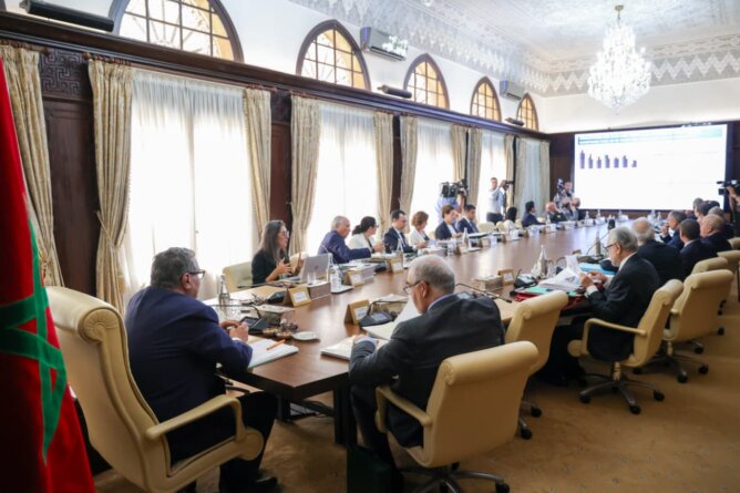 اجتماع رسمي في غرفة مزخرفة لمناقشة تقارير مالية مع علم المغرب وشاشة عرض