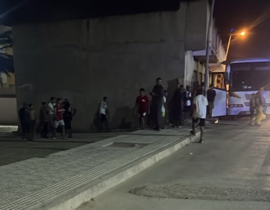 مجموعة أشخاص قرب حافلة بيضاء ليلاً في موقف حافلات قرب جدار رمادي