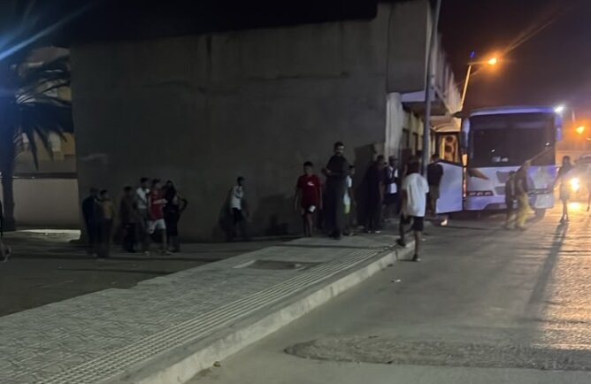 مجموعة أشخاص قرب حافلة بيضاء ليلاً في موقف حافلات قرب جدار رمادي
