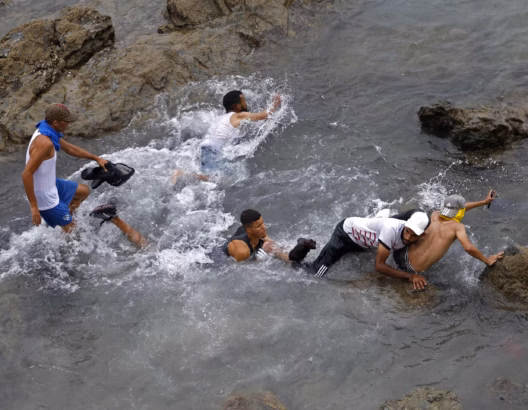 أشخاص يسبحون ويلعبون في مياه ضحلة قرب صخور وأحذية مبعثرة، صورة تعبيرية عن الهجرة