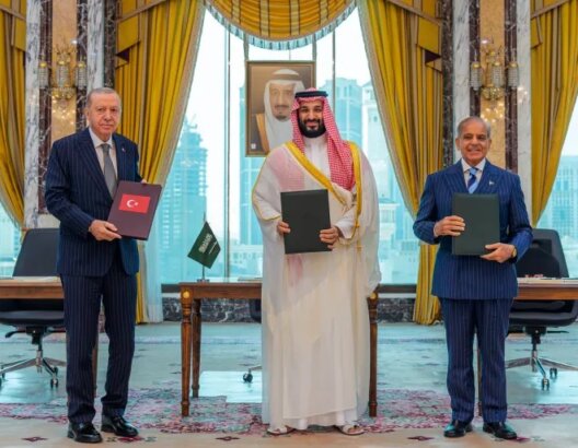 ثلاثة مسؤولين يحملون مجلدات أمام علمي السعودية وباكستان في غرفة رسمية