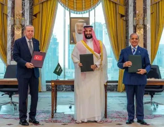 ثلاثة مسؤولين يحملون مجلدات أمام علمي السعودية وباكستان في غرفة رسمية