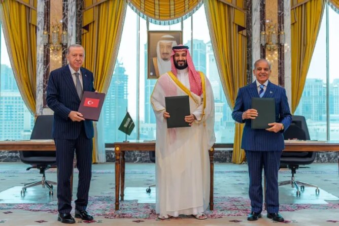 ثلاثة مسؤولين يحملون مجلدات أمام علمي السعودية وباكستان في غرفة رسمية