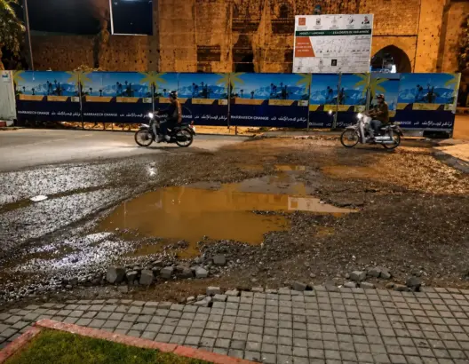طريق غير مهيأ بمراكش ليلاً مع بركة ماء ولوحات إعلانية ودراجتين ناريتين