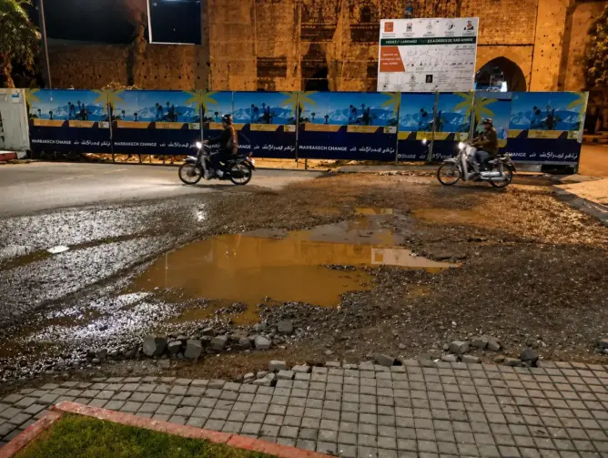 طريق غير مهيأ بمراكش ليلاً مع بركة ماء ولوحات إعلانية ودراجتين ناريتين