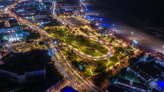 صورة جوية ليلية للواجهة البحرية بأكادير، تُظهر المساحات الخضراء والحدائق والطرق الملتفة بمحاذاة الشاطئ، إلى جانب مواقف السيارات والمنشآت السياحية المنتشرة على الساحل.